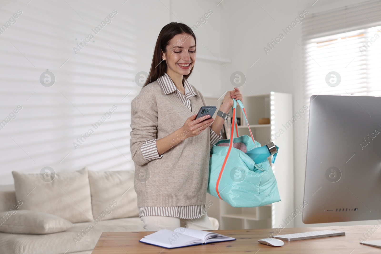 Photo of Happy woman with gym bag and smartphone at desk indoors