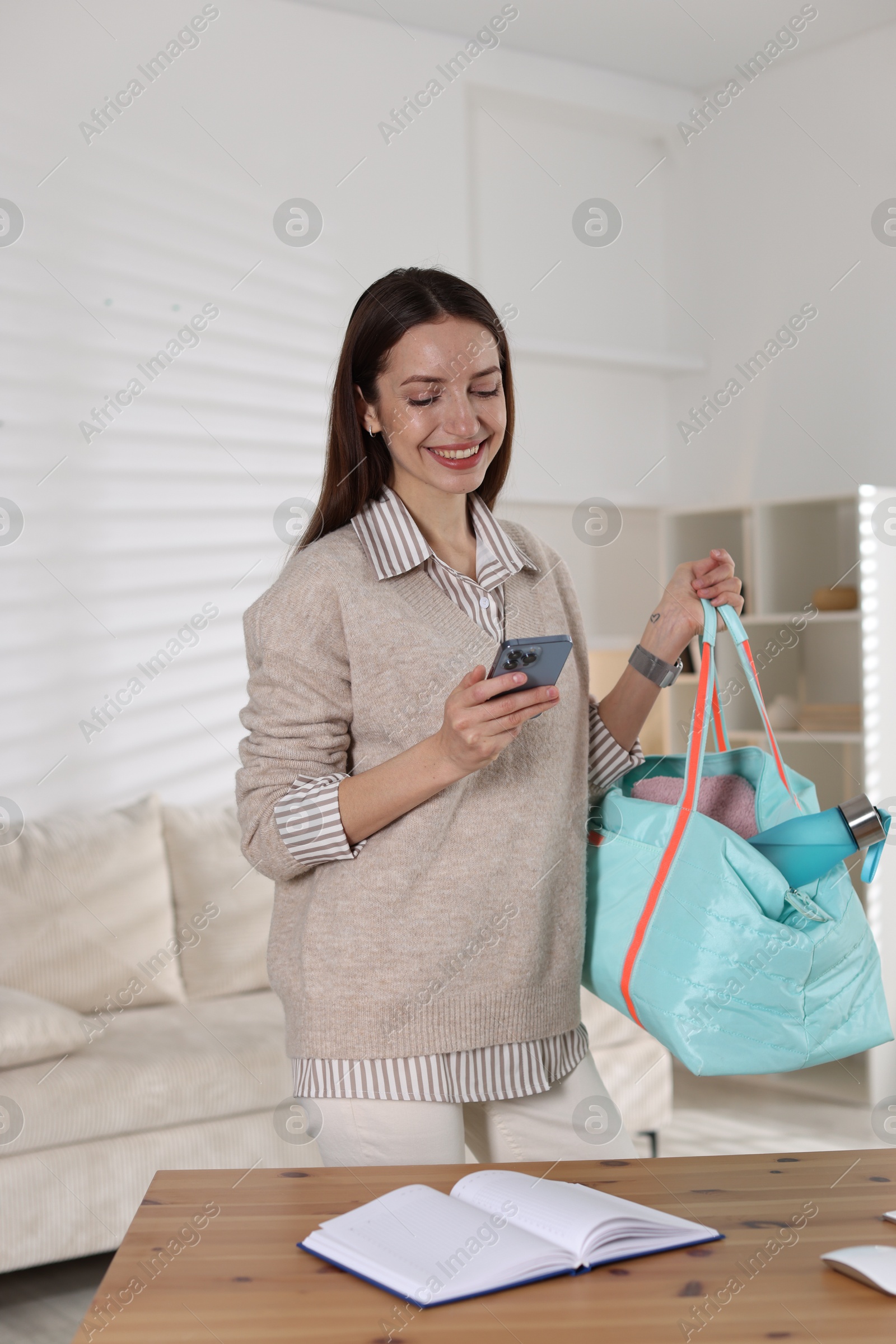 Photo of Happy woman with gym bag and smartphone at desk indoors