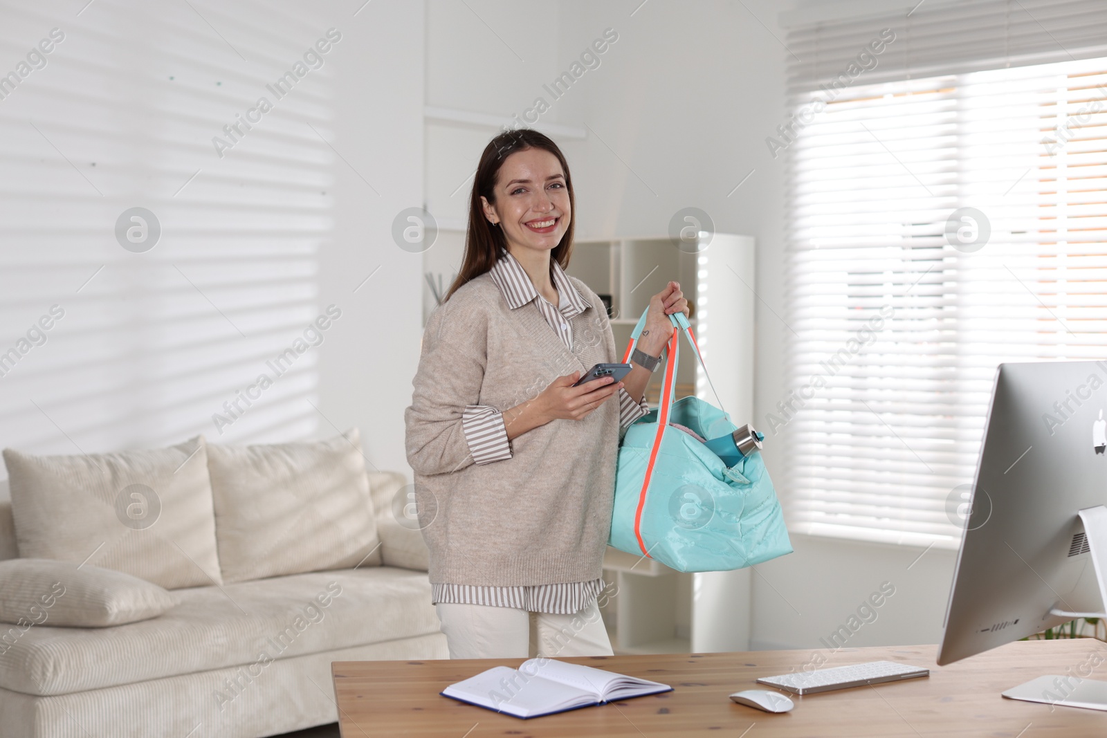 Photo of Happy woman with gym bag and smartphone at desk indoors