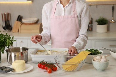 Photo of Woman spreading bechamel sauce onto spinach lasagna at marble table in kitchen, closeup