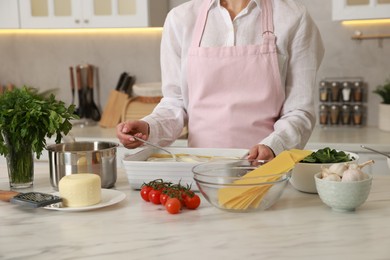 Photo of Woman spreading bechamel sauce onto spinach lasagna at marble table in kitchen, closeup