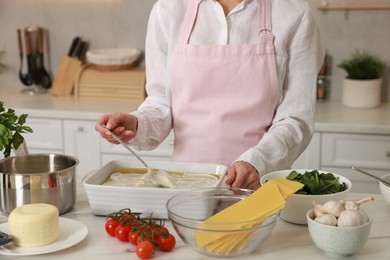 Photo of Woman spreading bechamel sauce onto spinach lasagna at marble table in kitchen, closeup