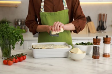 Photo of Woman grating cheese onto spinach lasagna at marble table indoors, closeup