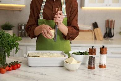 Photo of Woman grating cheese onto spinach lasagna at marble table indoors, closeup