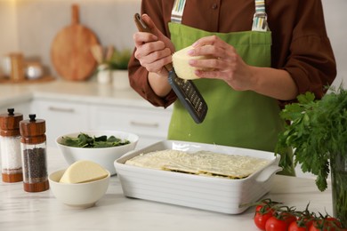 Photo of Woman grating cheese onto spinach lasagna at marble table indoors, closeup