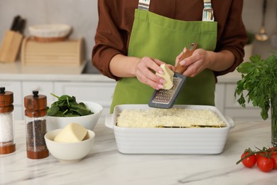 Photo of Woman grating cheese onto spinach lasagna at marble table indoors, closeup