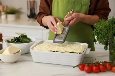 Photo of Woman grating cheese onto spinach lasagna at marble table indoors, closeup