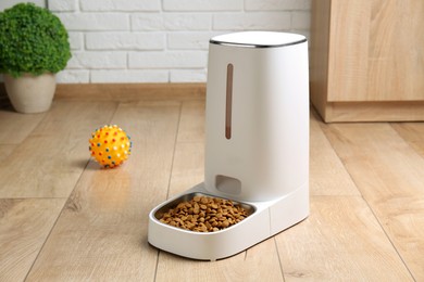 Photo of Dry pet food in automatic feeder and toy on floor indoors