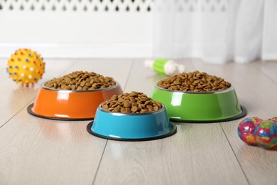 Photo of Dry pet food in feeding bowls and toys on floor indoors