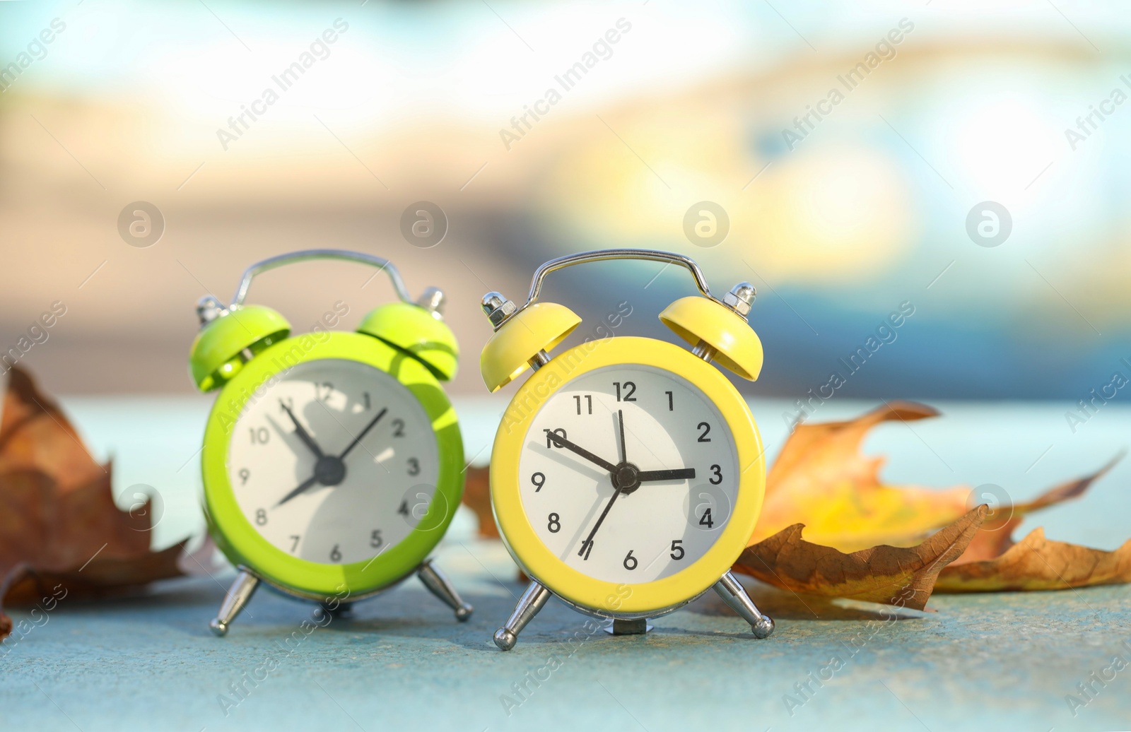 Photo of Autumn time. Alarm clocks and golden leaves on light surface outdoors, closeup