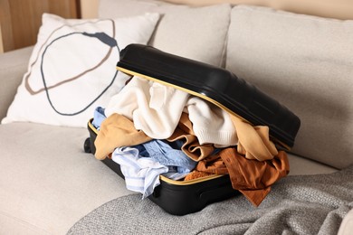 Photo of Open suitcase full of different messy clothes on sofa indoors
