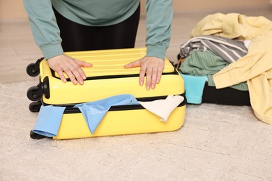 Photo of Woman trying to close suitcase full of different messy clothes indoors, closeup