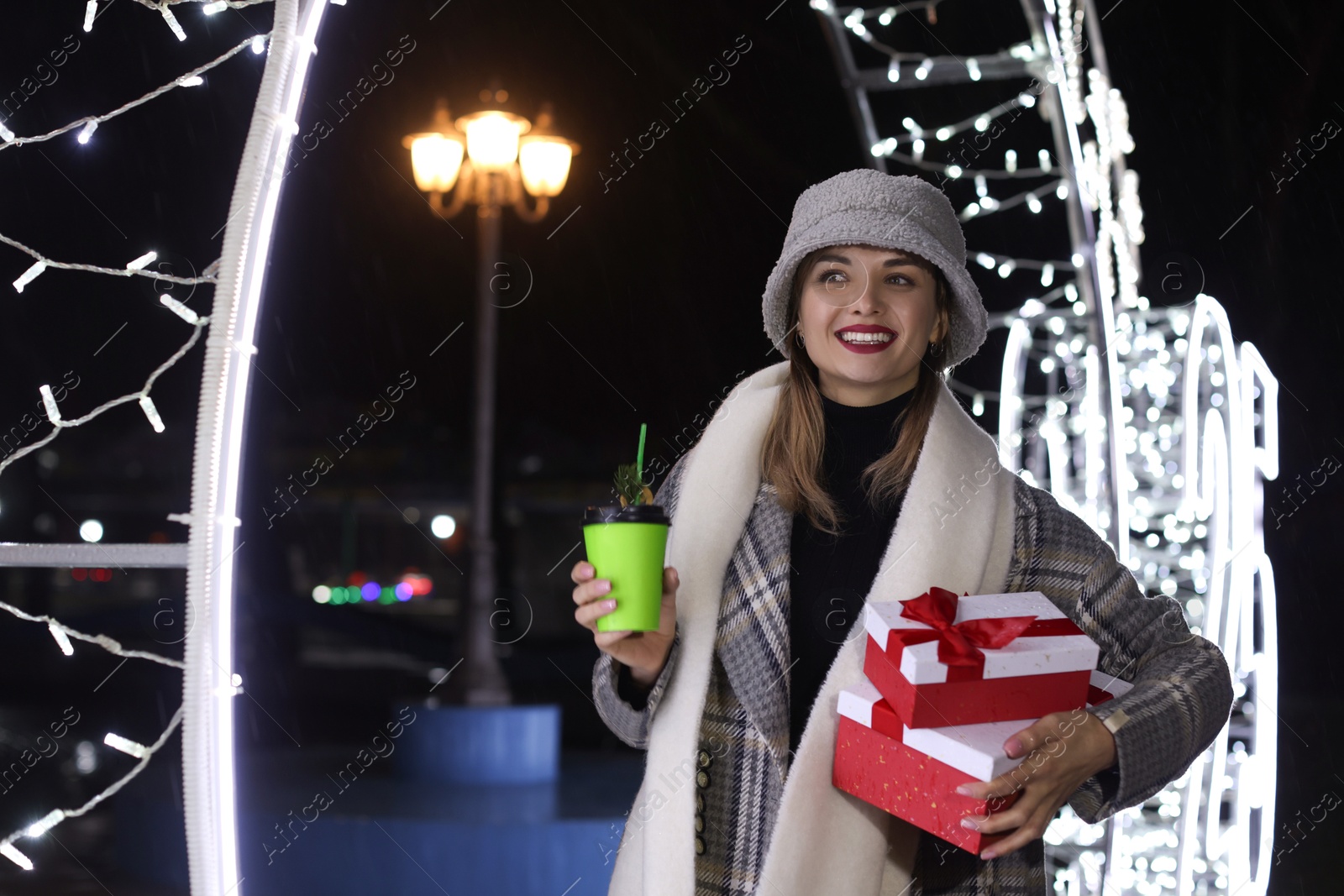 Photo of Happy woman with gifts and paper cup near Christmas decoration at night outdoors