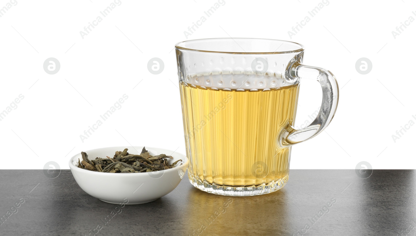 Photo of Refreshing green tea in cup and dry leaves on grey textured table against white background