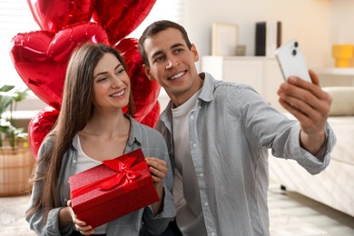 Photo of Lovely couple with gift box taking selfie near heart shaped balloons indoors. Valentine's day celebration