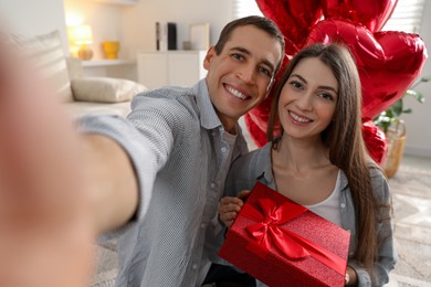 Photo of Lovely couple with gift box taking selfie near heart shaped balloons indoors. Valentine's day celebration