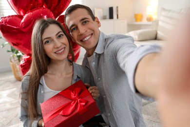 Photo of Lovely couple with gift box taking selfie near heart shaped balloons indoors. Valentine's day celebration