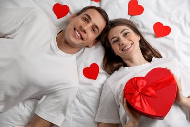 Photo of Lovely couple lying on bed with paper hearts and gift box, top view. Valentine's day celebration