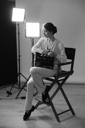 Photo of Beautiful woman with clapperboard sitting in director's chair in studio. Black and white effect