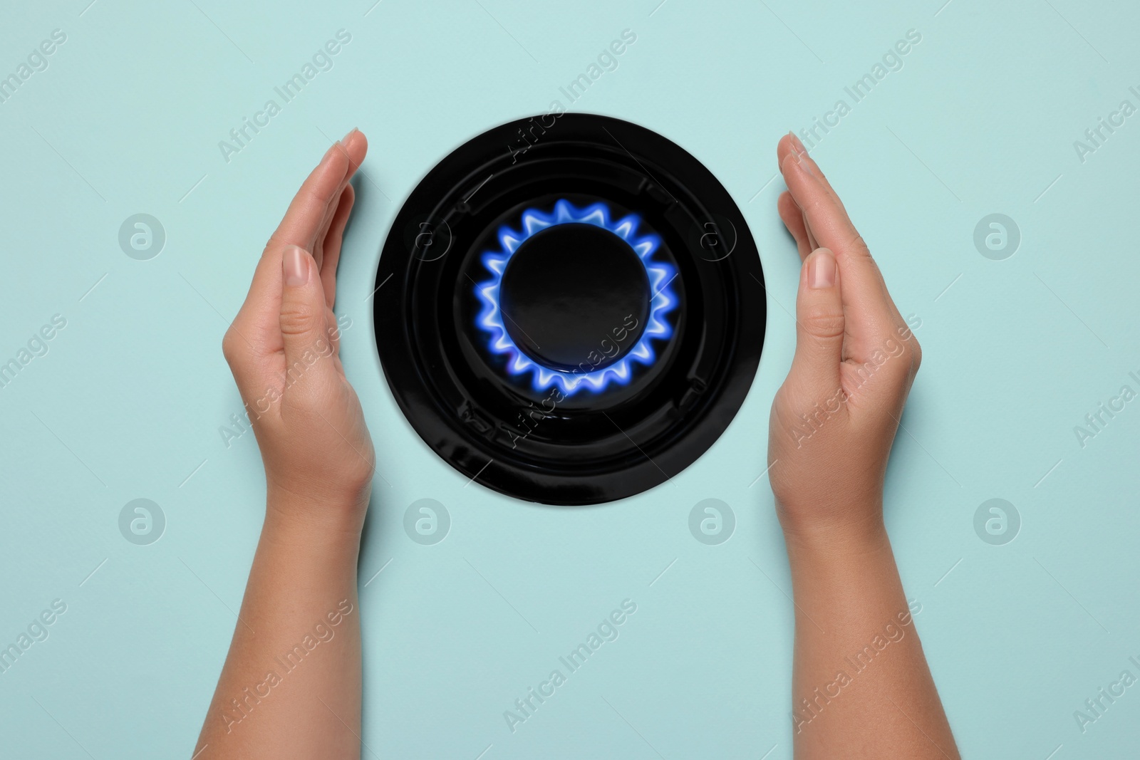 Image of Woman protecting gas burner with blue flame on light blue background, top view