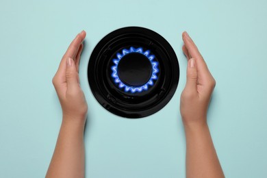 Image of Woman protecting gas burner with blue flame on light blue background, top view