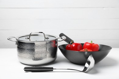 Photo of Ladle, pot and wok with vegetables on white table. Cooking utensils