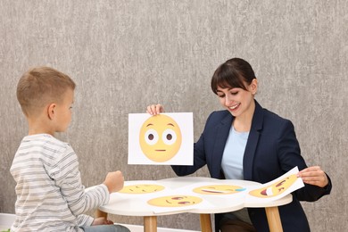 Photo of Autism therapy. Little boy choosing emoticon at table with smiling psychologist in mental health center