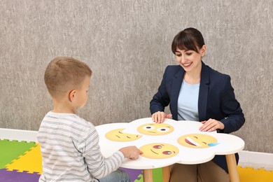 Photo of Autism therapy. Little boy choosing emoticon at table with smiling psychologist in mental health center, back view