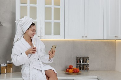 Photo of Spa day. Beautiful woman with face mask and glass of juice using smartphone in kitchen