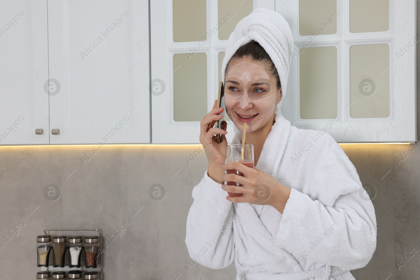 Photo of Spa day. Beautiful woman with face mask and glass of juice talking on smartphone in kitchen