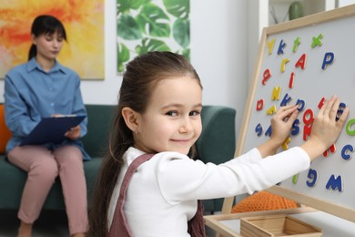 Photo of Girl assembling letters on magnetic board while psychologist taking notes indoors, selective focus