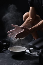 Photo of Man clapping hands with talcum powder above bowl before training on black background, closeup