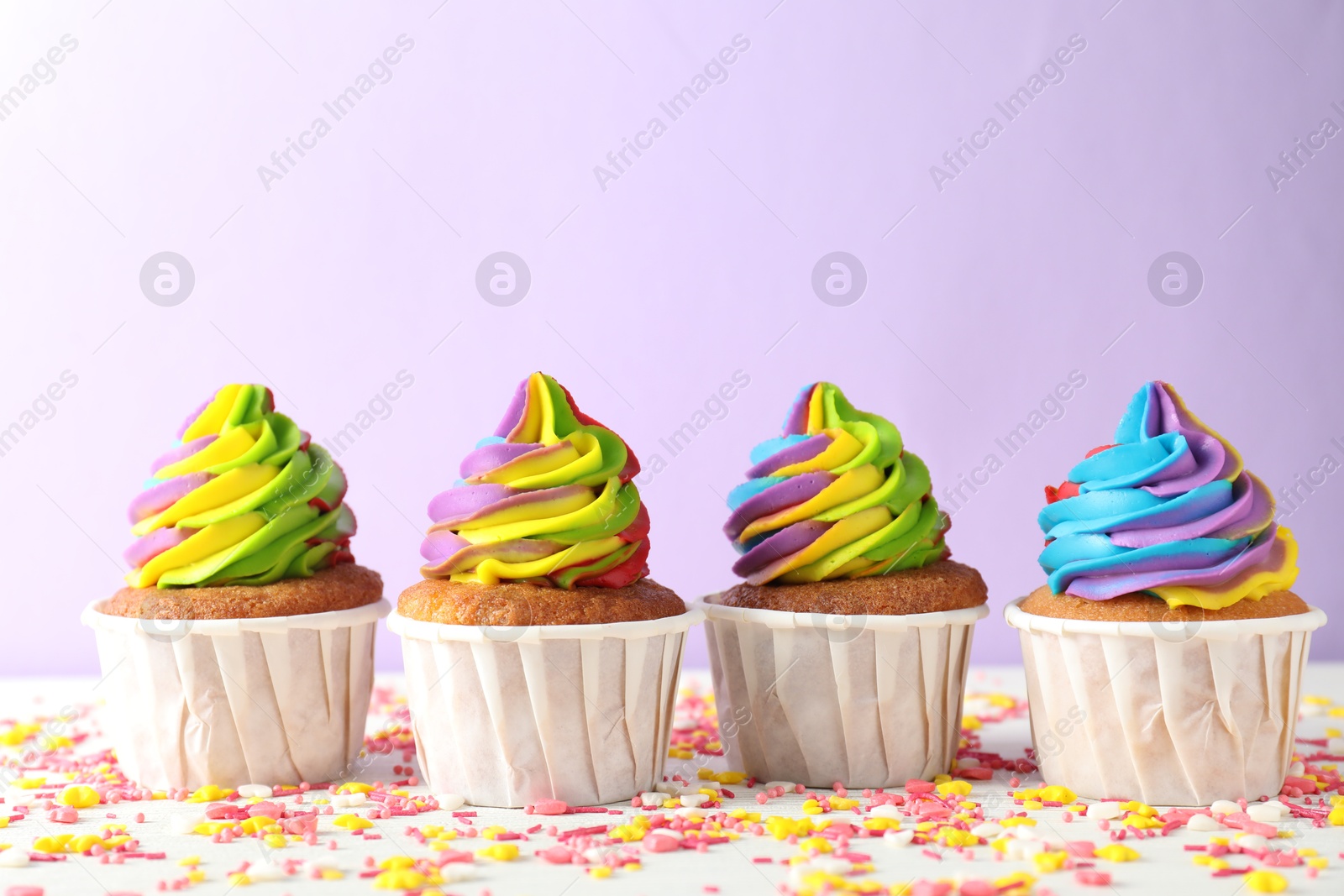 Photo of Delicious cupcakes with colorful cream and sprinkles on white table against violet background, closeup