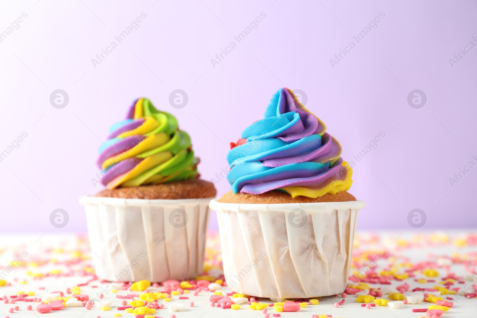 Photo of Delicious cupcakes with colorful cream and sprinkles on white table against violet background, closeup