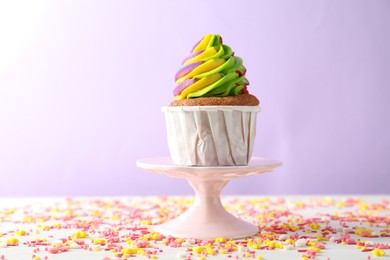 Photo of One delicious cupcake with colorful cream and sprinkles on white table against violet background, closeup