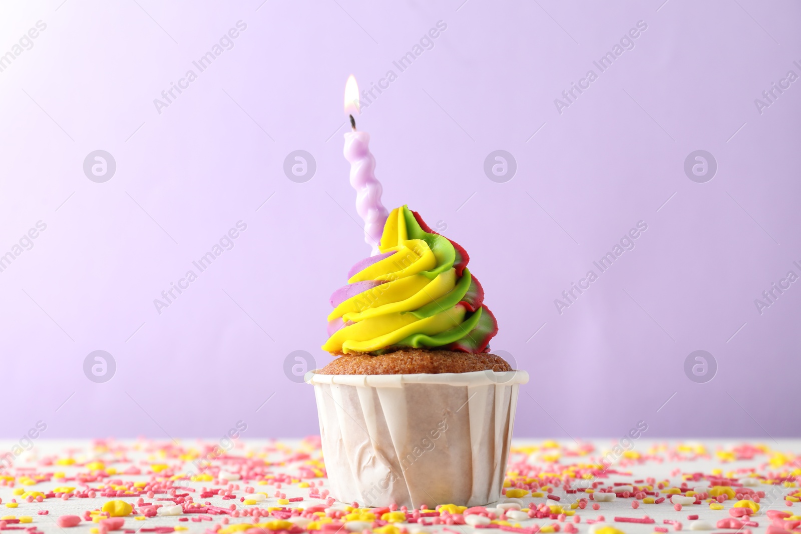 Photo of One delicious cupcake with colorful cream, burning candle and sprinkles on white table against violet background, closeup