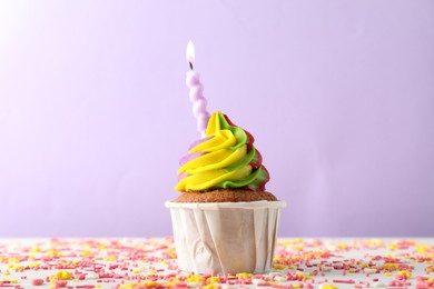 Photo of One delicious cupcake with colorful cream, burning candle and sprinkles on white table against violet background, closeup