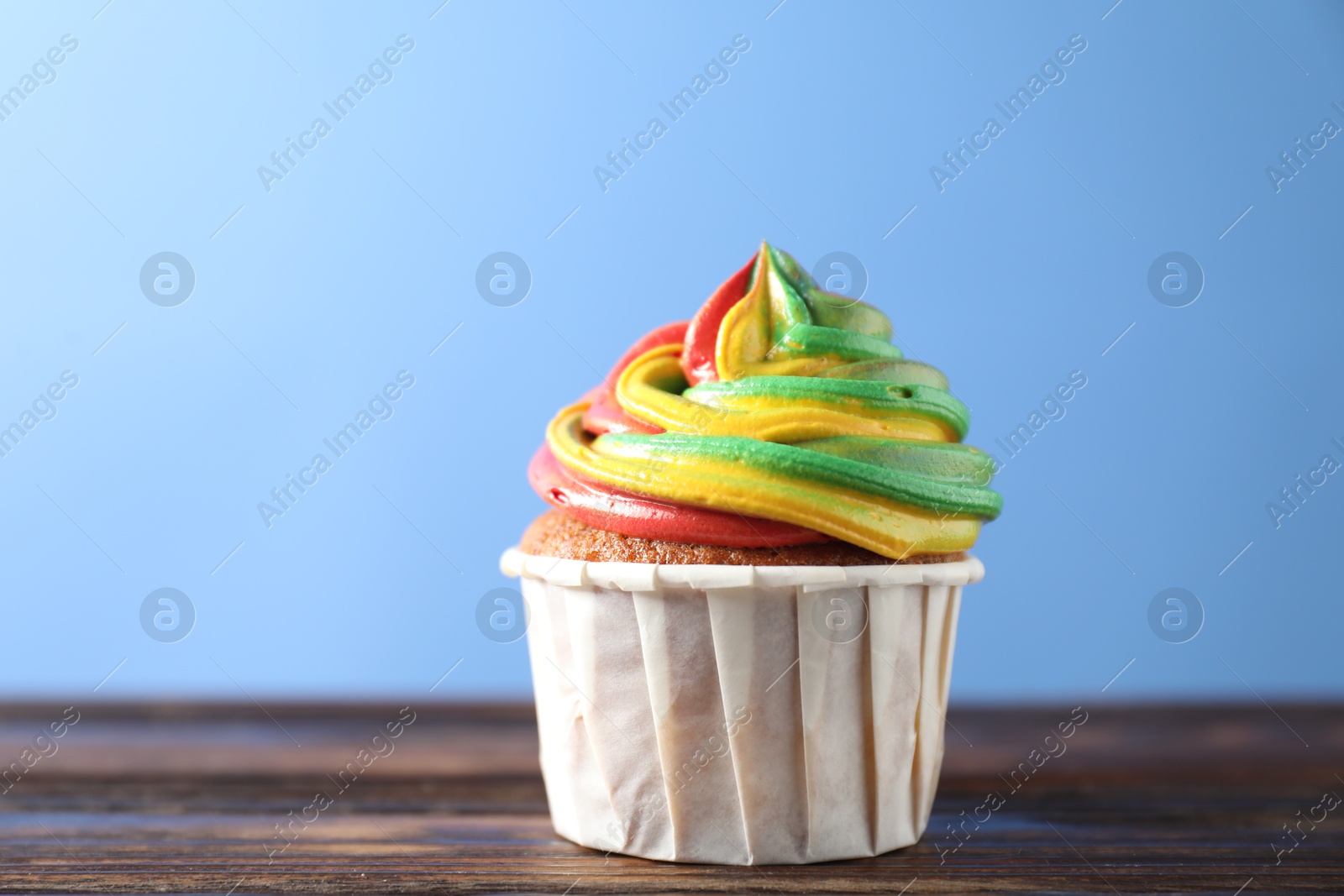 Photo of Delicious cupcake with colorful cream on wooden table against blue background, closeup