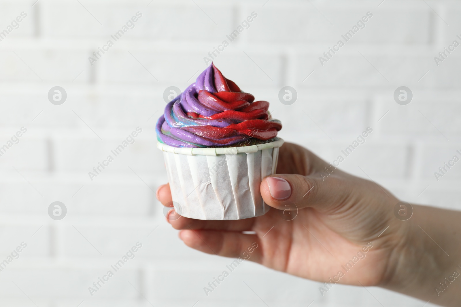 Photo of Woman holding delicious cupcake with colorful cream against white brick wall, closeup