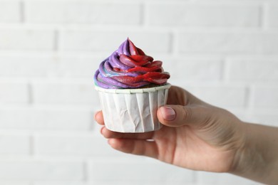 Photo of Woman holding delicious cupcake with colorful cream against white brick wall, closeup