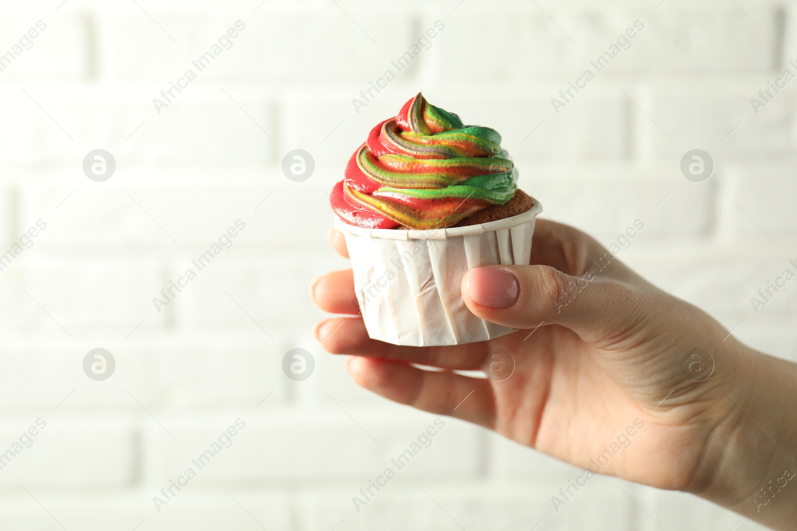 Photo of Woman holding delicious cupcake with colorful cream against white brick wall, closeup