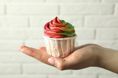 Photo of Woman holding delicious cupcake with colorful cream against white brick wall, closeup