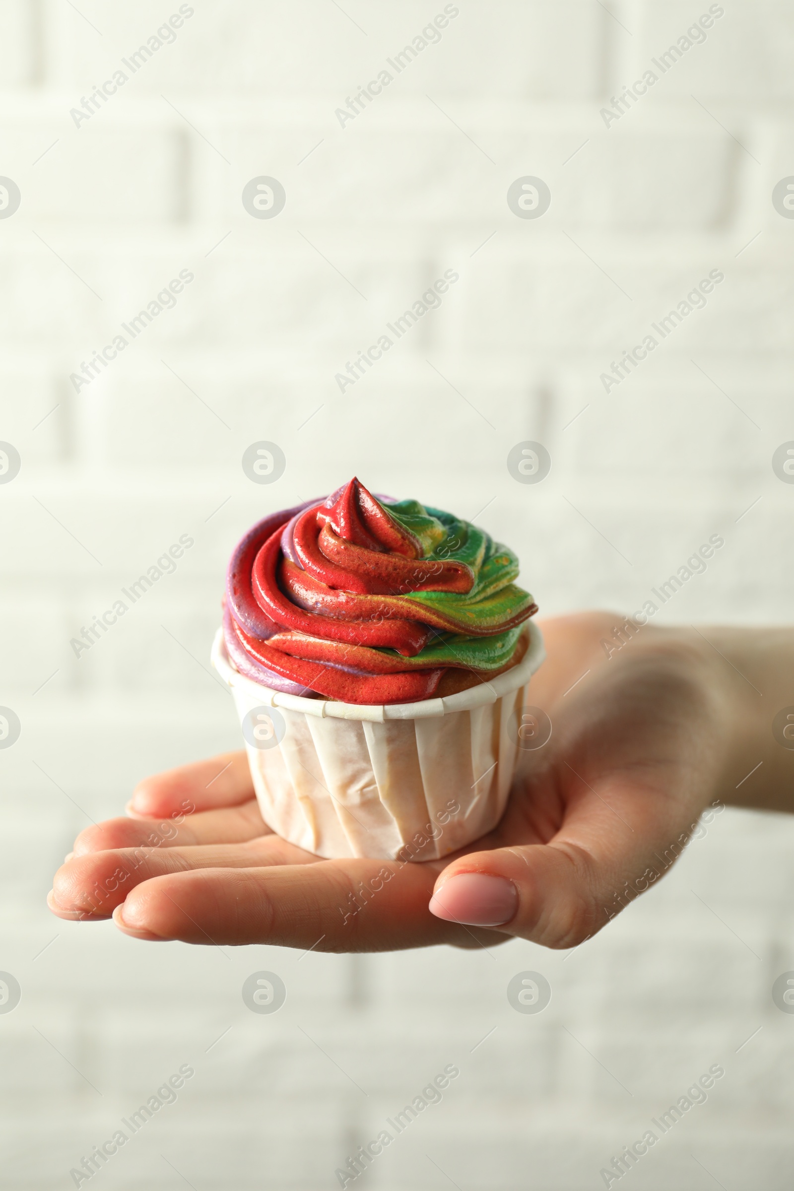 Photo of Woman holding delicious cupcake with colorful cream against white brick wall, closeup. Space for text