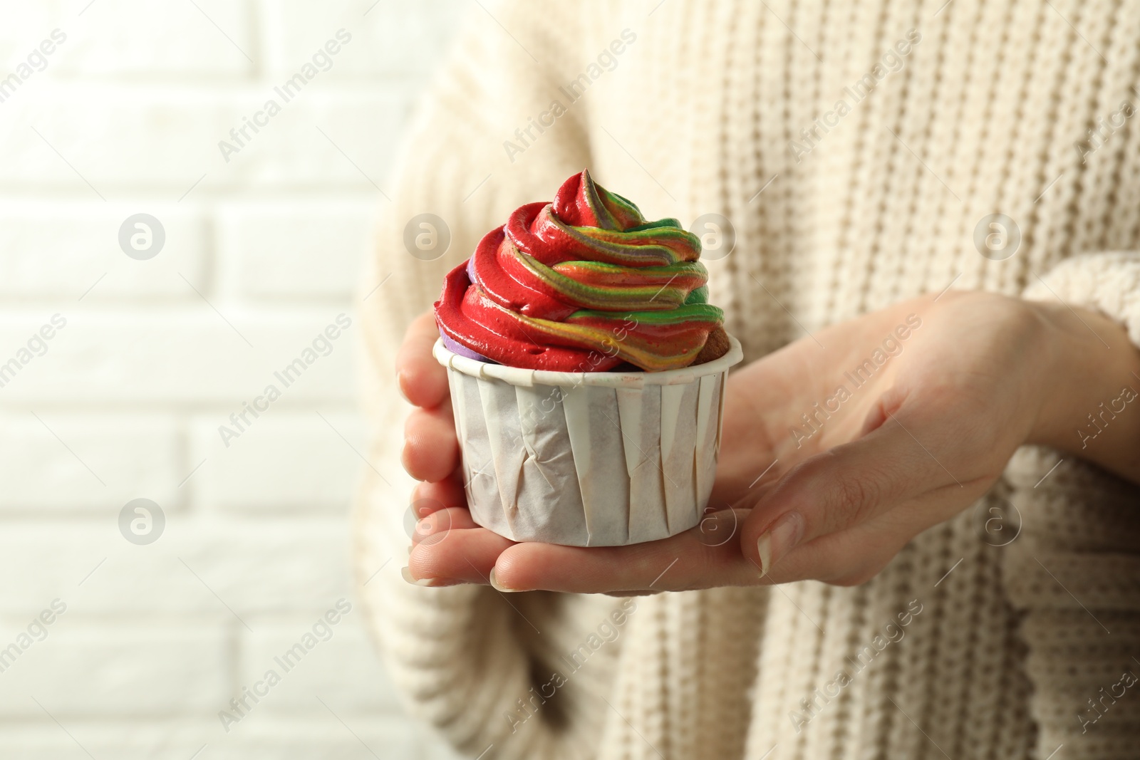 Photo of Woman holding delicious cupcake with colorful cream against white brick wall, closeup