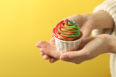 Photo of Woman holding delicious cupcake with colorful cream on yellow background, closeup. Space for text