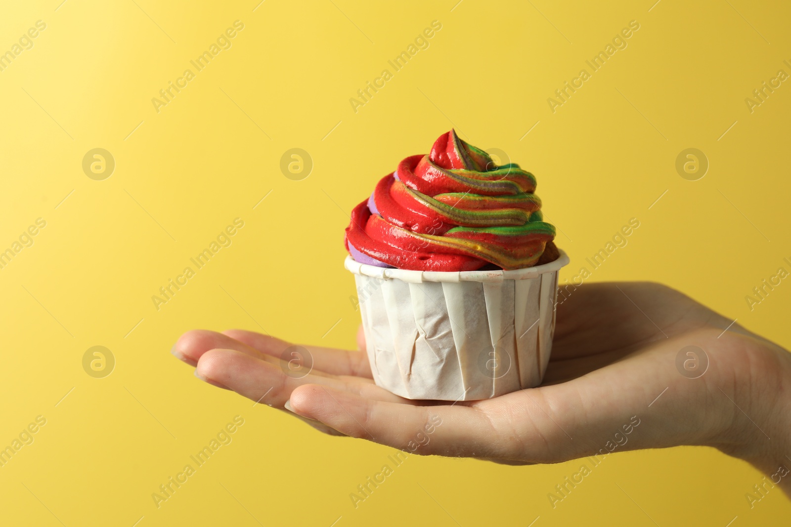 Photo of Woman holding delicious cupcake with colorful cream on yellow background, closeup