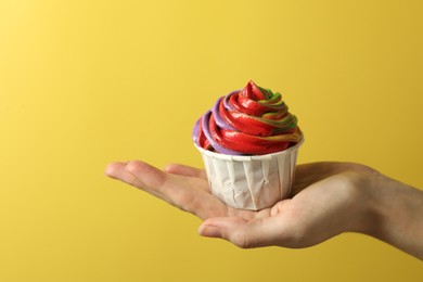 Photo of Woman holding delicious cupcake with colorful cream on yellow background, closeup. Space for text