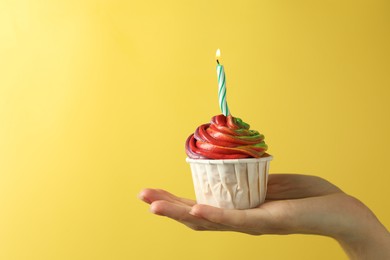 Photo of Woman holding delicious cupcake with colorful cream and burning candle on yellow background, closeup. Space for text