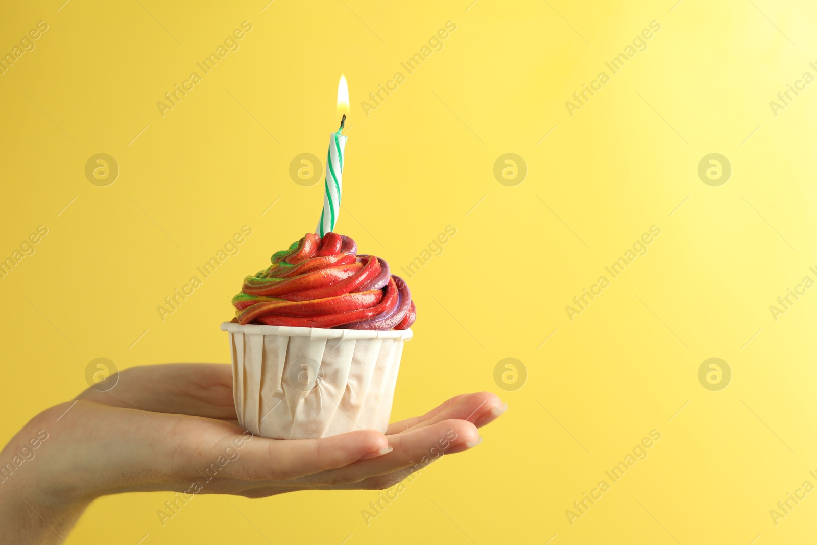 Photo of Woman holding delicious cupcake with colorful cream and burning candle on yellow background, closeup. Space for text