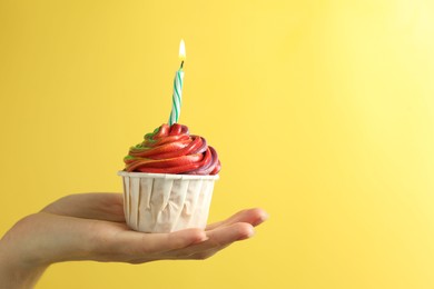 Photo of Woman holding delicious cupcake with colorful cream and burning candle on yellow background, closeup. Space for text